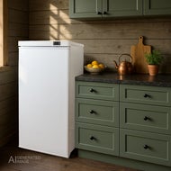 AI-genererad bild av ett Kylskåp Sunwind Ventus Low Energy 160, 230V, Vit står bredvid gröna köksskåp i ett rum med träpanel på väggarna. På bänkskivan finns en skål med citroner, en kopparfärgad vattenkokare, en skärbräda och en krukväxt. Dagsljus faller in från ett fönster till vänster.