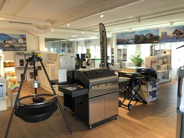 En butik med grillar och utomhusprodukter från Sunwind utställda på ett välbelyst trägolv. I förgrunden står en bålpanna och i bakgrunden syns olika grillmodeller, tillbehör och informationsskyltar. Ljusa väggar och stora fönster släpper in naturligt ljus som ger en inbjudande atmosfär