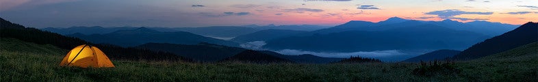 Ett tält lyser varmt i skymningen på en gräsbevuxen höjd med vid utsikt över blå bergssiluetter och dimfyllda dalar under en färgstark kvällshimmel