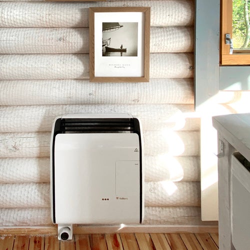 En vit väggmonterad gasolkamin är installerad på en ljus timmervägg i en stuga. Ovanför värmaren hänger en träram med ett svartvitt fotografi. Till höger syns ett fönster som släpper in solljus, och golvet består av ljusa trägolvbrädor