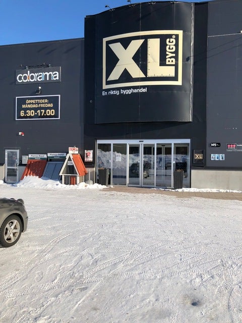 Entrén till byggvaruhuset XL Bygg under en solig vinterdag. Marken är täckt av snö och skyltar med öppettider och varumärken, som Colorama, syns på den mörkgrå fasaden