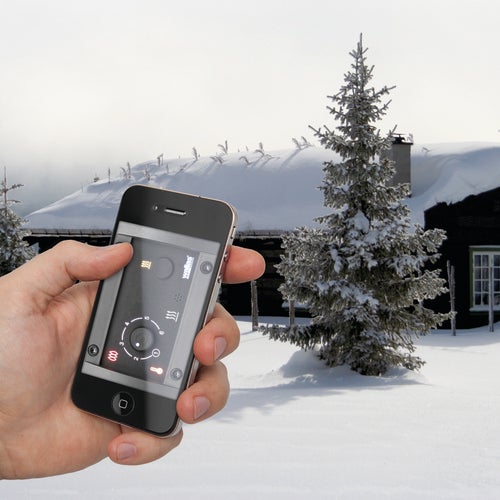 Hand som håller en smartphone med en styrningsapp, framför en snötäckt stuga i vinterlandskap