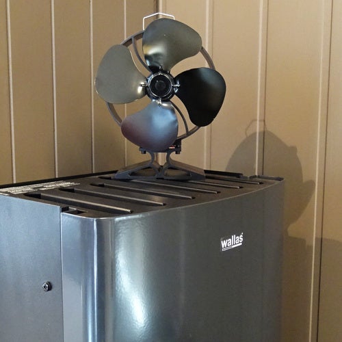 En svart värmefläkt med etiketten ”Wallas” står mot en beige träpanelvägg. Ovanpå fläkten finns en kaminfläkt Neat med fyra metallblad