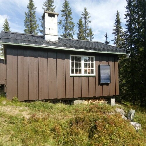 En brun trästuga med vit skorsten och ett fönster på gaveln. Bredvid fönstret sitter en solventilator AirPlus monterad på väggen. Stugan ligger på en gräsbevuxen sluttning omgiven av granskog under en klarblå himmel