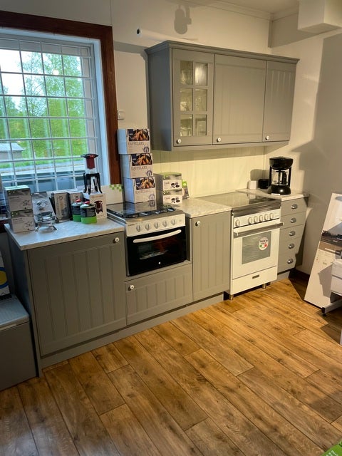 Ett utställningskök med grå köksskåp, två spisar och en kaffebryggare på bänken. Golvet är ett trägolv och ett stort fönster släpper in naturligt ljus