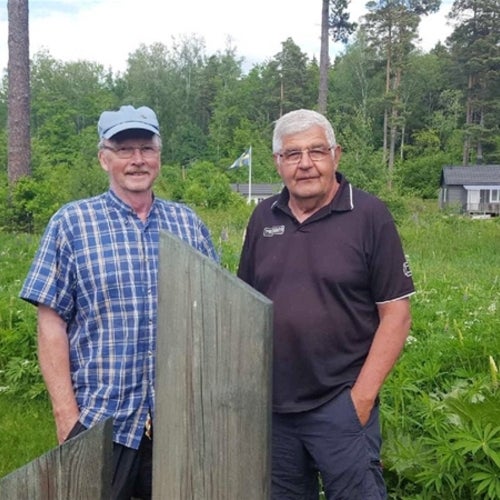Två äldre män står utomhus i en grönskande trädgård med träd, blommor och små stugor i bakgrunden. De ler mot kameran, den ena bär blå keps och rutig skjorta, den andra har en mörk pikétröja
