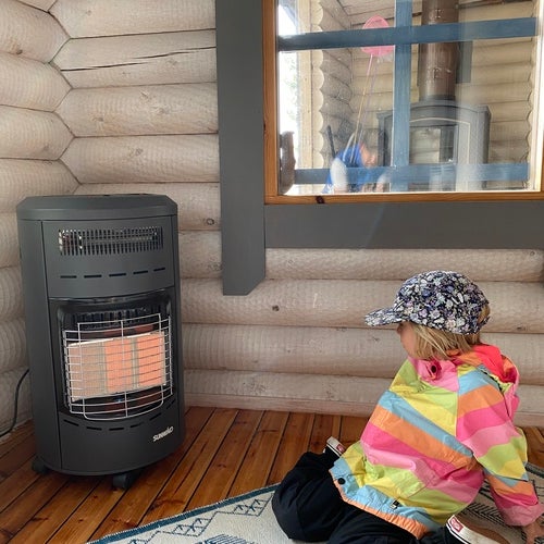 Ett barn i färgglad randig jacka och keps sitter på golvet i en timrad stuga framför en svart gasolkamin Fan-tastic med synlig låga. Barnet tittar mot värmaren, och bakom syns ett fönster med reflektion av en skorsten. Stämningen är varm och hemtrevlig
