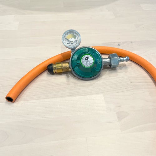 En gasregulator med tryckmätare och ansluten orange slang ligger på ett ljust trägolv. Utrustningen används för att koppla gas till exempelvis grillar eller spisar