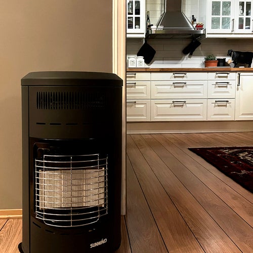 En svart gasolkamin står placerad i ett hem med trägolv, framför en öppning mot ett kök med vita köksluckor, rostfri spisfläkt och köksredskap hängande på väggen