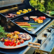 Gasolgrill med kött, korv, grönsaker och majs som grillas, med färdiggrillad mat och färgglada grönsaker upplagda på ett fat bredvid.