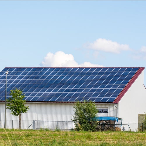 En vit byggnad med ett stort sadeltak täckt av blå solpaneler står på en grästäckt tomt. Några träd och en vedhög syns framför huset, och himlen är klarblå med några vita moln