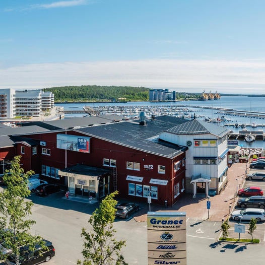 En röd handelsbyggnad vid en småbåtshamn en solig dag. Framför byggnaden står en skylt med företagsnamn, och i bakgrunden syns havet, båtar och moderna bostadshus längs kajen