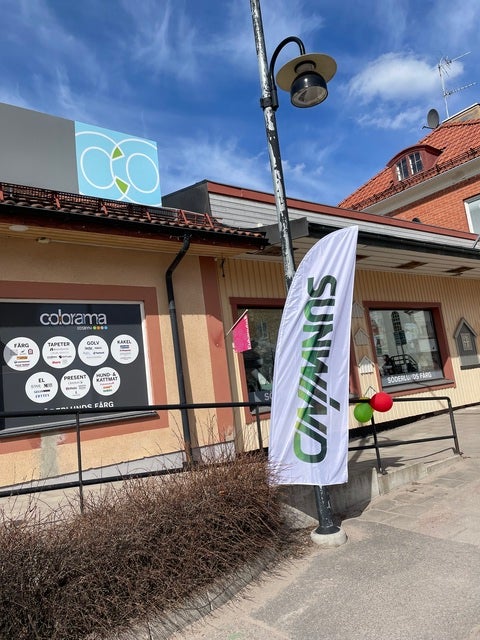 En gata med en byggnad som har skyltar för “Colorama” och “Sunwind”. Framför byggnaden står en vit flagga med Sunwinds logotyp, och himlen är klarblå med några moln