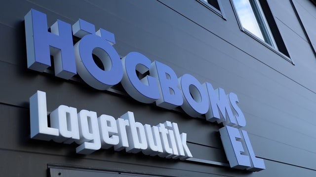 En byggnad med en skylt som visar texten ”Högboms El Lagerbutik” i stora bokstäver på en mörk fasad. Ovanför skylten syns några fönster som reflekterar himlen