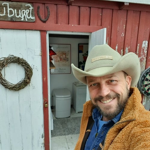 En man med cowboyhatt och brun fleecejacka ler mot kameran framför en röd stuga med vit dörr. Inne i stugan syns en toalett och tavlor på väggen. På dörren hänger en krans, och ovanför står en skylt med texten "Utburet" samt en hästsko