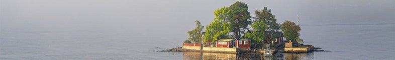 En liten ö med röda stugor, träd och en brygga ligger stilla i dimmig sjö. Vattnet är lugnt och omgivningen har ett mjukt, disigt morgonljus