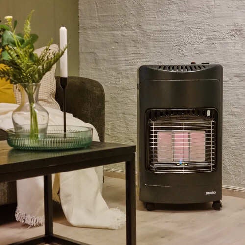 En gasolkamin Windflame från Sunwind står i ett vardagsrum mot en grå vägg. Framför kaminen finns ett soffbord med en vas med gröna växter och ett tänt stearinljus. I bakgrunden syns en soffa med en vit filt